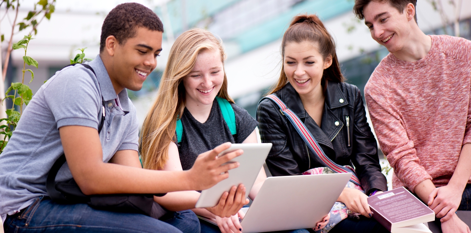 a group of four students smiling at a laptop and a tablet